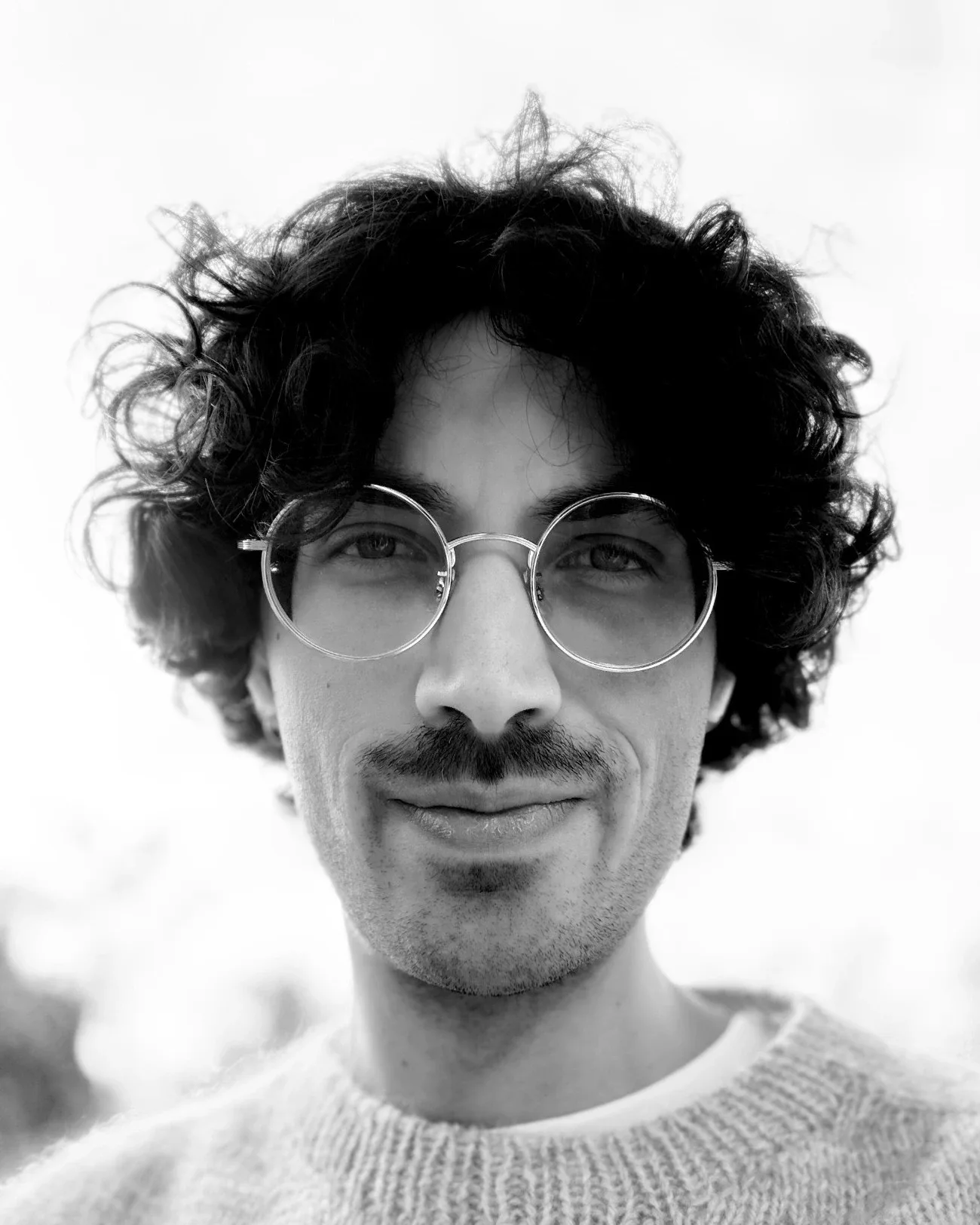 Schwarzweiß-Porträt von Raphael Mayer mit lockigen Haaren, runder Brille und Strickpullover vor hellem Himmel